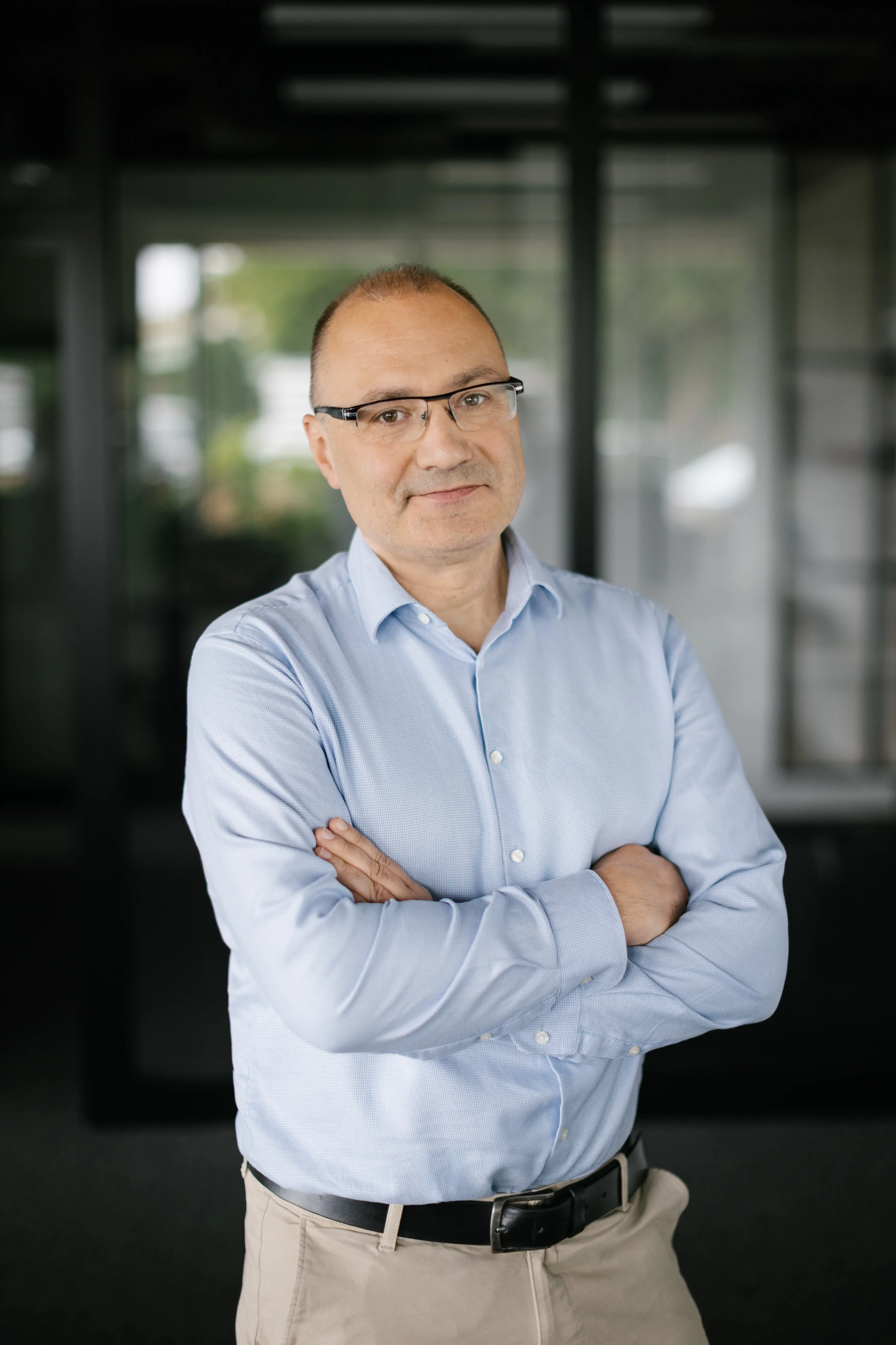 Mężczyzna w średnim wieku z okularami stoi w pomieszczeniu ze skrzyżowanymi rękami, ubrany w jasnoniebieską koszulę zapinaną na guziki i beżowe spodnie, patrząc w kamerę z lekkim uśmiechem.