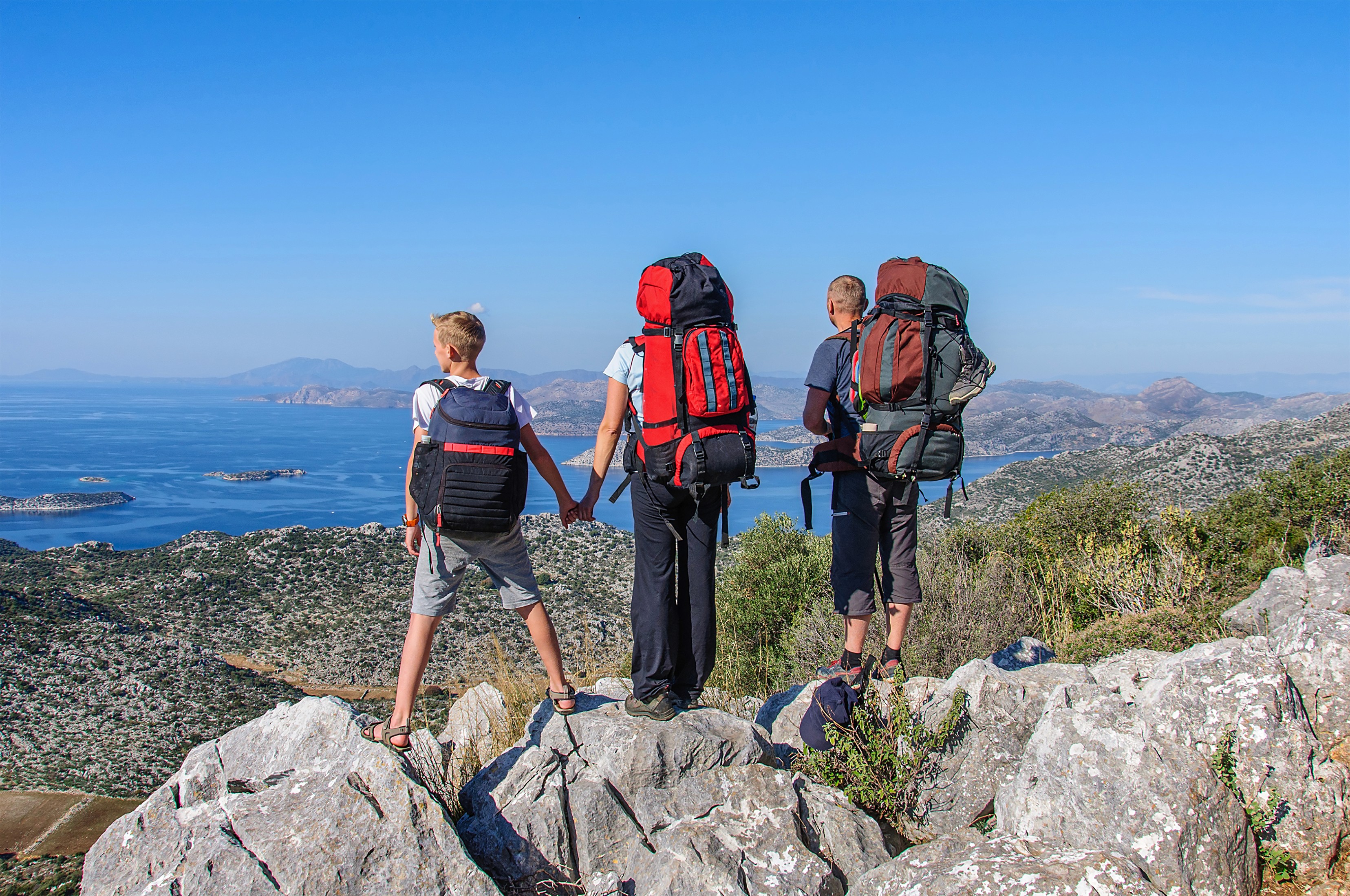 Three hikers with backpacks standing on a rocky cliff overlooking the sea.