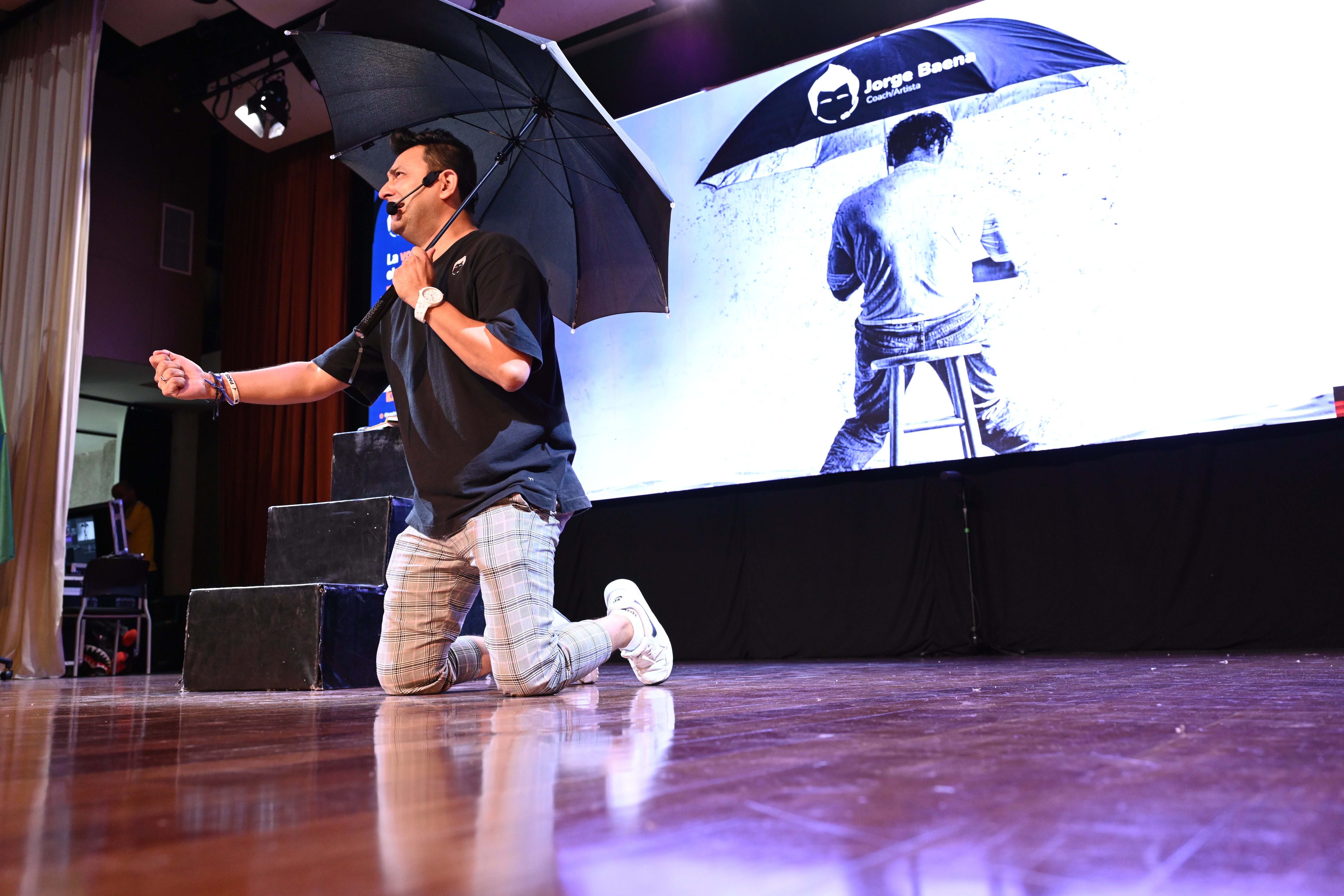 Jorge Baena arrodillado en el escenario sosteniendo un paraguas negro durante una conferencia, con una gran pantalla detrás que muestra la misma imagen del paraguas como parte de su presentación visual.