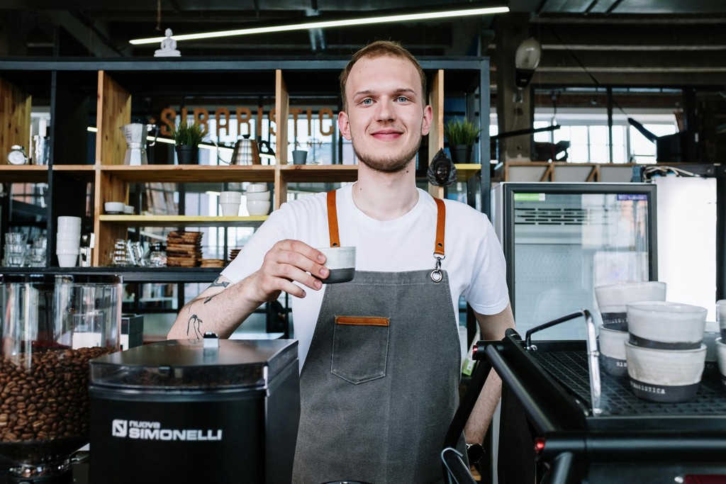 A smiling newcomer to Canada wearing an apron while working as a barista behind the counter of a local cafe, representing popular part-time survival jobs in the service industry for international students and immigrants.