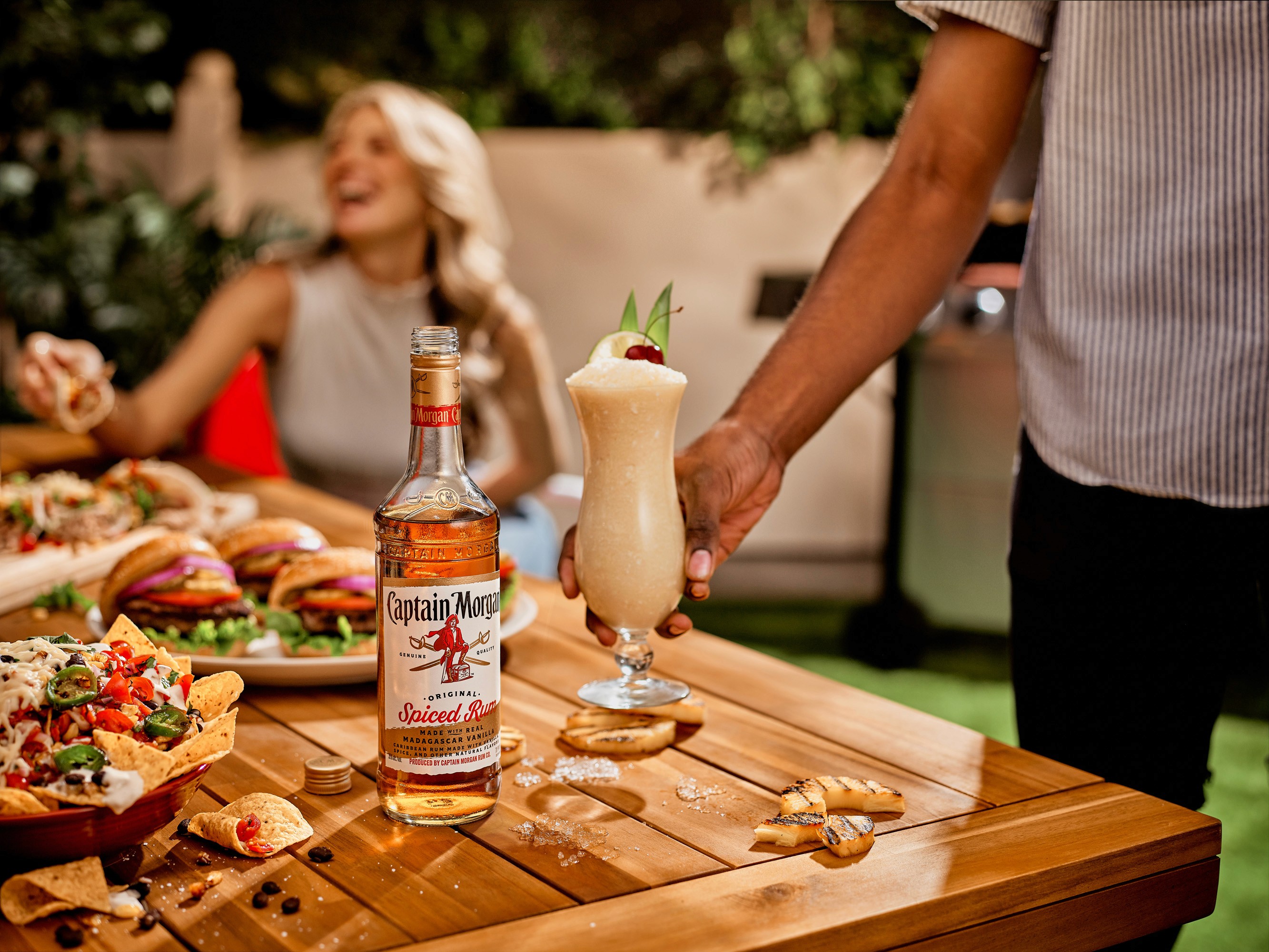 Sunny garden party with a person serving a frothy pina colada next to a bottle of Captain Morgan Spiced Rum, with joyful guests and a table full of delicious burgers and nachos in the background.