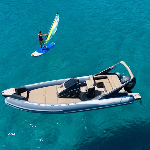 An inflatable motorboat on clear blue water with a person windsurfing nearby.