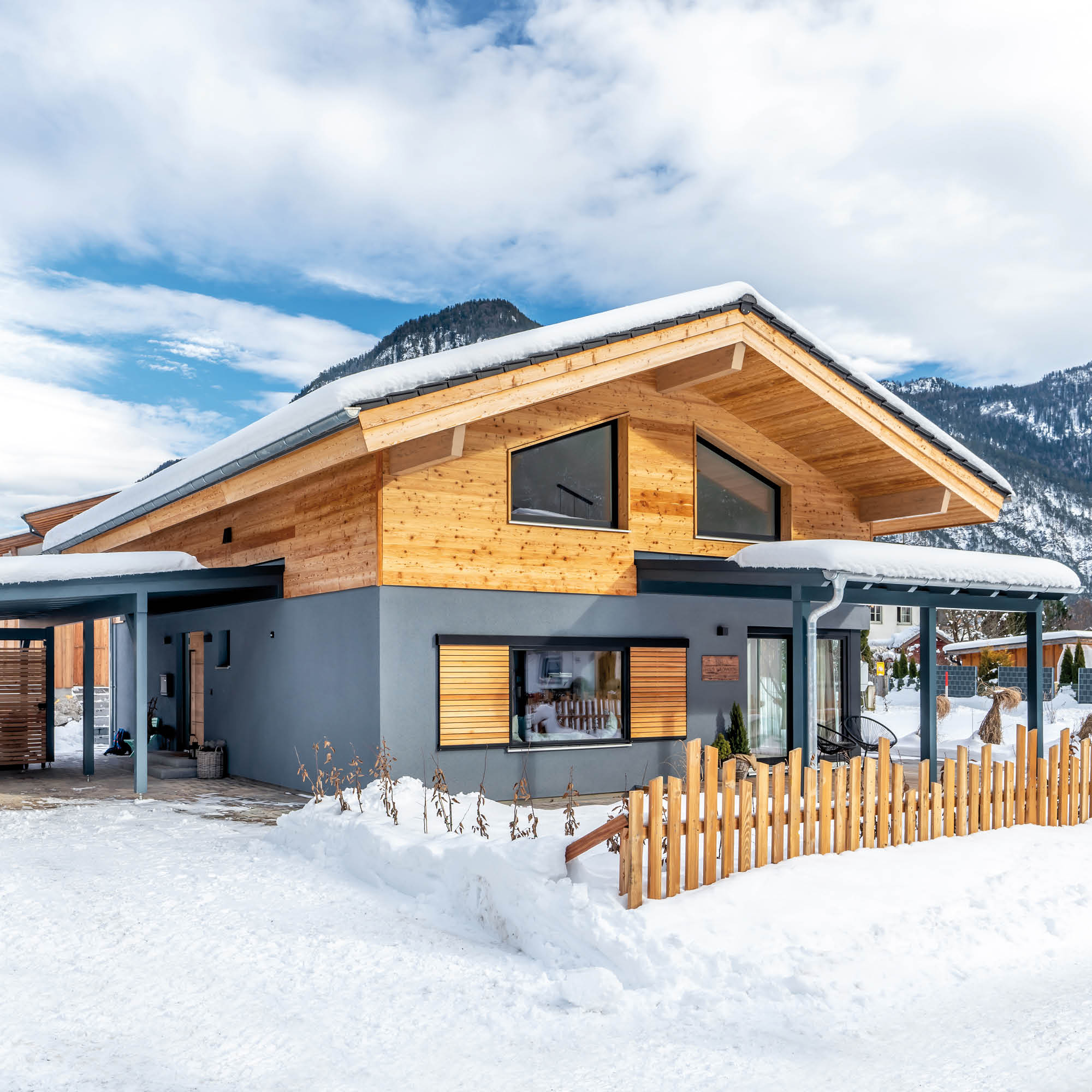 Modernes Holzhaus mit grauem Putz und Schneebedecktem Dach, umgeben von Schnee und Bergen.
