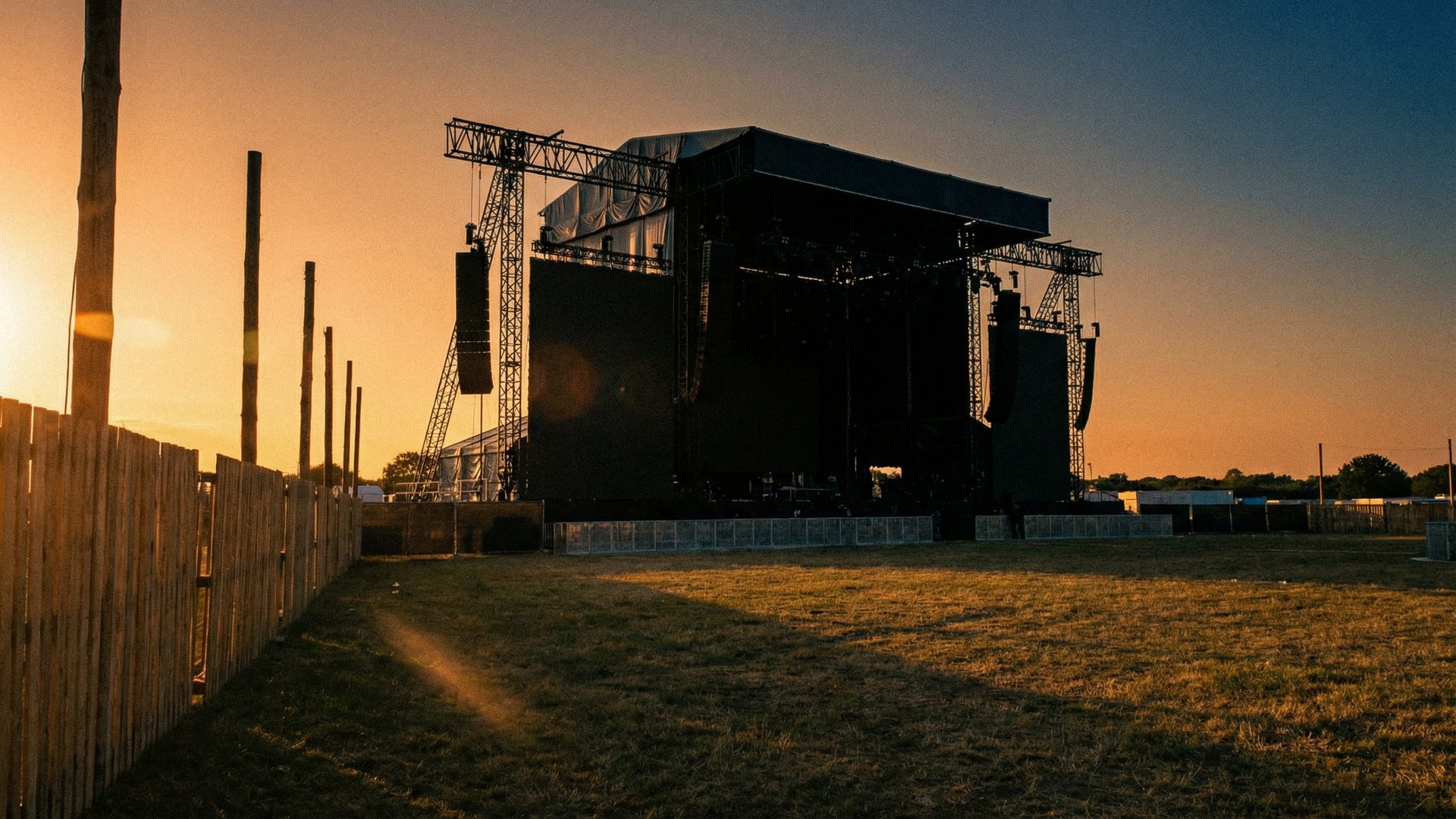 An empty outdoor festival grounds at golden hour, with a massive main stage structure silhouetted against a warm amber and deep blue sky, festival fencing and empty grass field in the foreground.