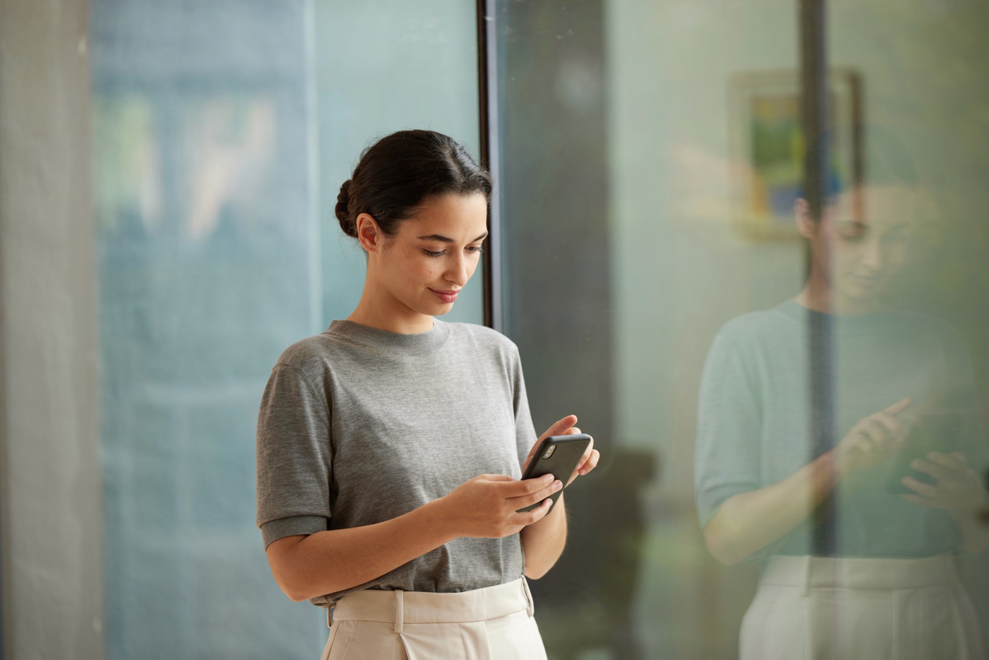 A young woman in a grey t-shirt looking at her smartphone while standing near glass doors in a modern building