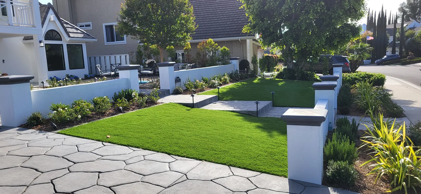 Broad paved paths and low steps cross a green front garden bordered by white walls and flowering plants.