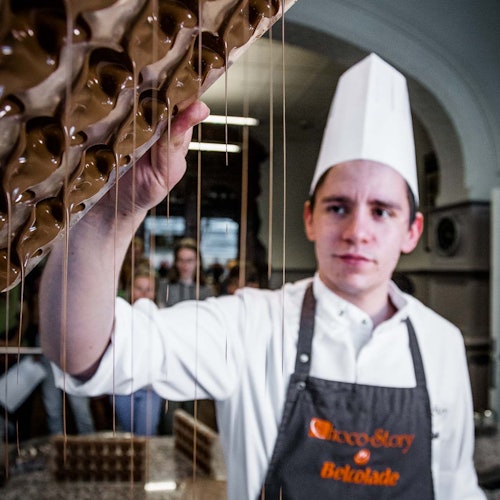 Egy szakács fehér kalapban és kötényben folyékony csokoládét önt ki egy öntőformából egy konyhában, miközben mások a háttérben figyelik.