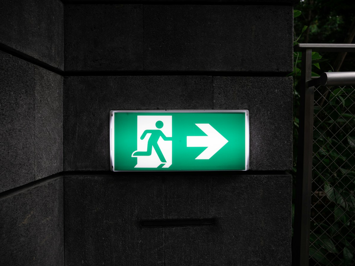 An illuminated green running-man exit sign against a dark architectural wall