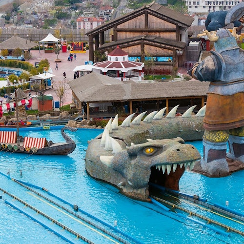 Outdoor na liwasan ng aliwan na may malaking dragon-themed na water ride, mga bangka, mga gusaling gawa sa kahoy, at mga taong naglalakad sa likuran.