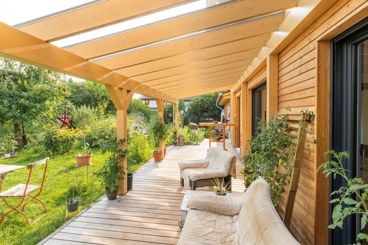 Holzterrassenüberdachung mit Sitzmöbeln, Pflanzen und Gartenmöbeln vor einem Holzhaus im Grünen.
