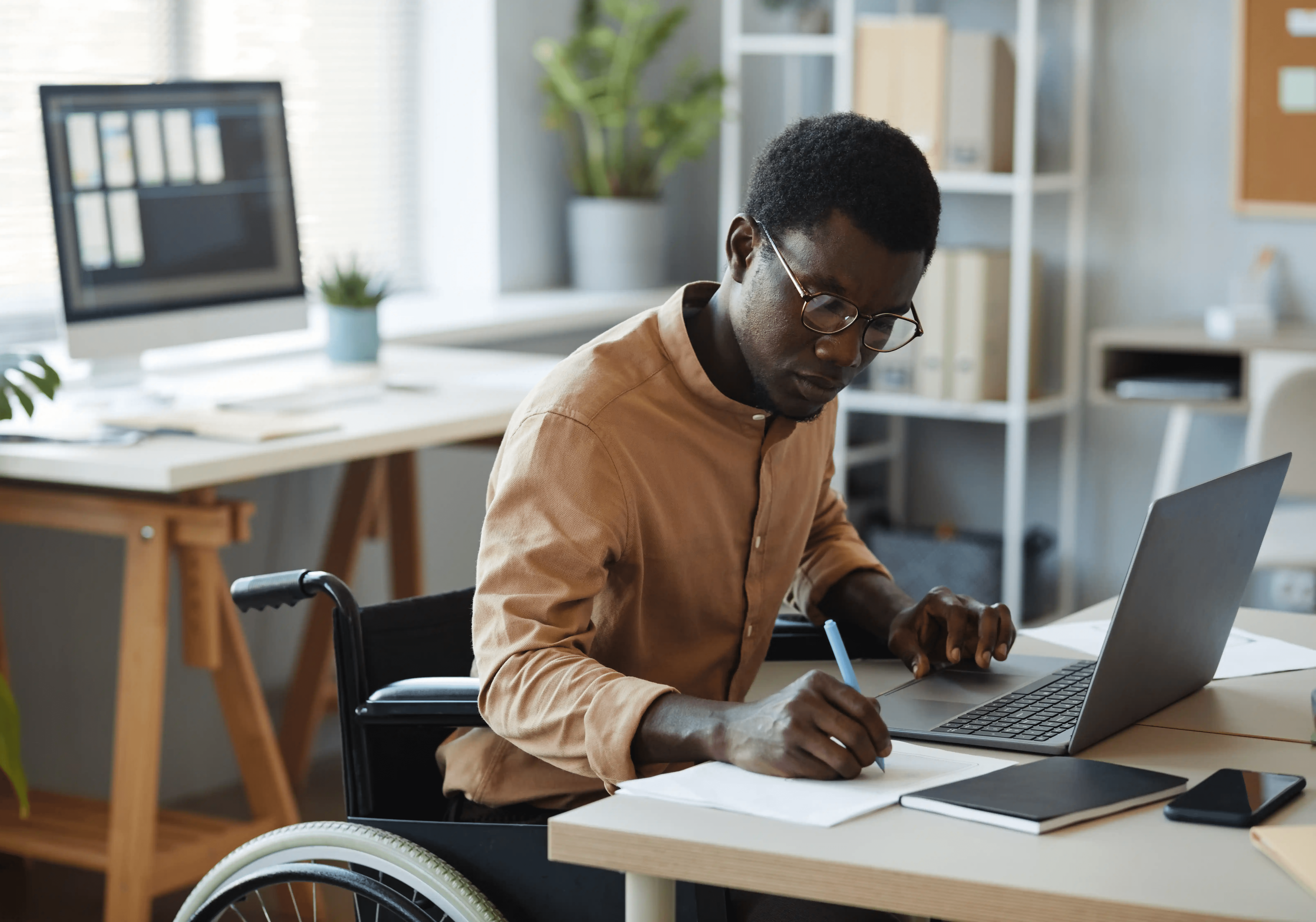 An office employee in a wheelchair writing notes on paper while working at a laptop desk, illustrating steps workers should take to document accommodation requests and protect their legal rights