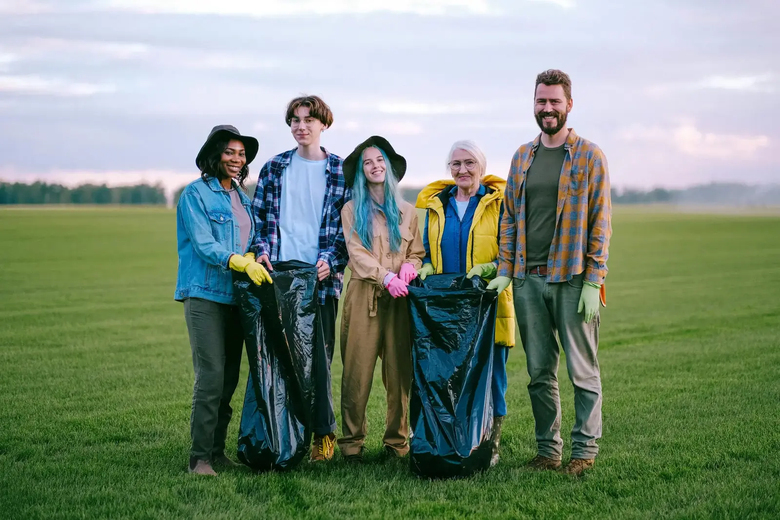 Group of people of various ages participating in a nature cleanup, holding trash bags and wearing clothing appropriate for outdoor tasks.