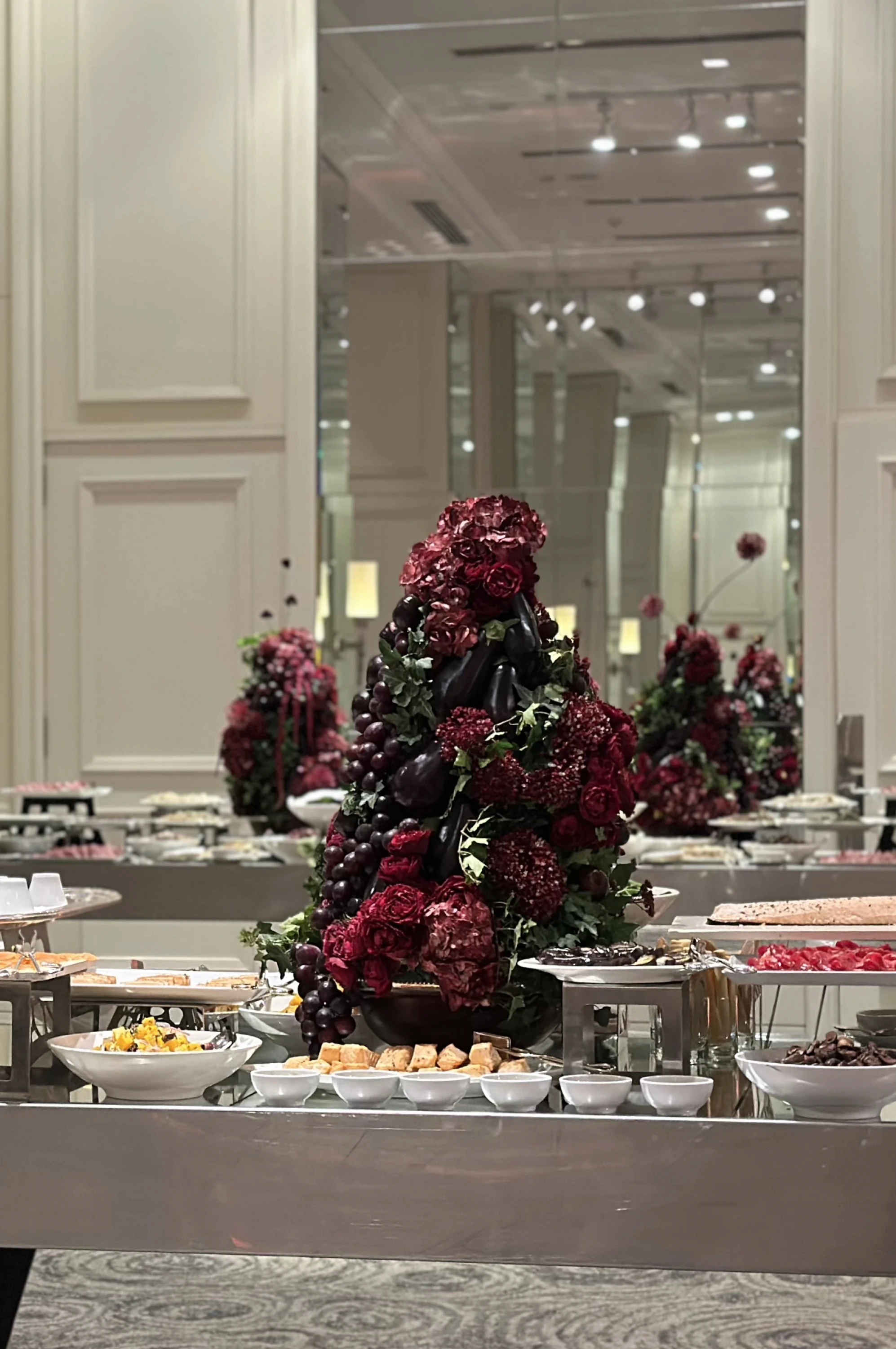 Estación de comida con imponente centro de mesa barroco de orquídeas y frutas frente a buffet en salón elegante.