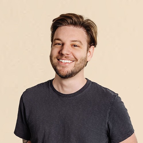Smiling man with short hair and light facial hair wearing a dark T-shirt, standing against a neutral background.