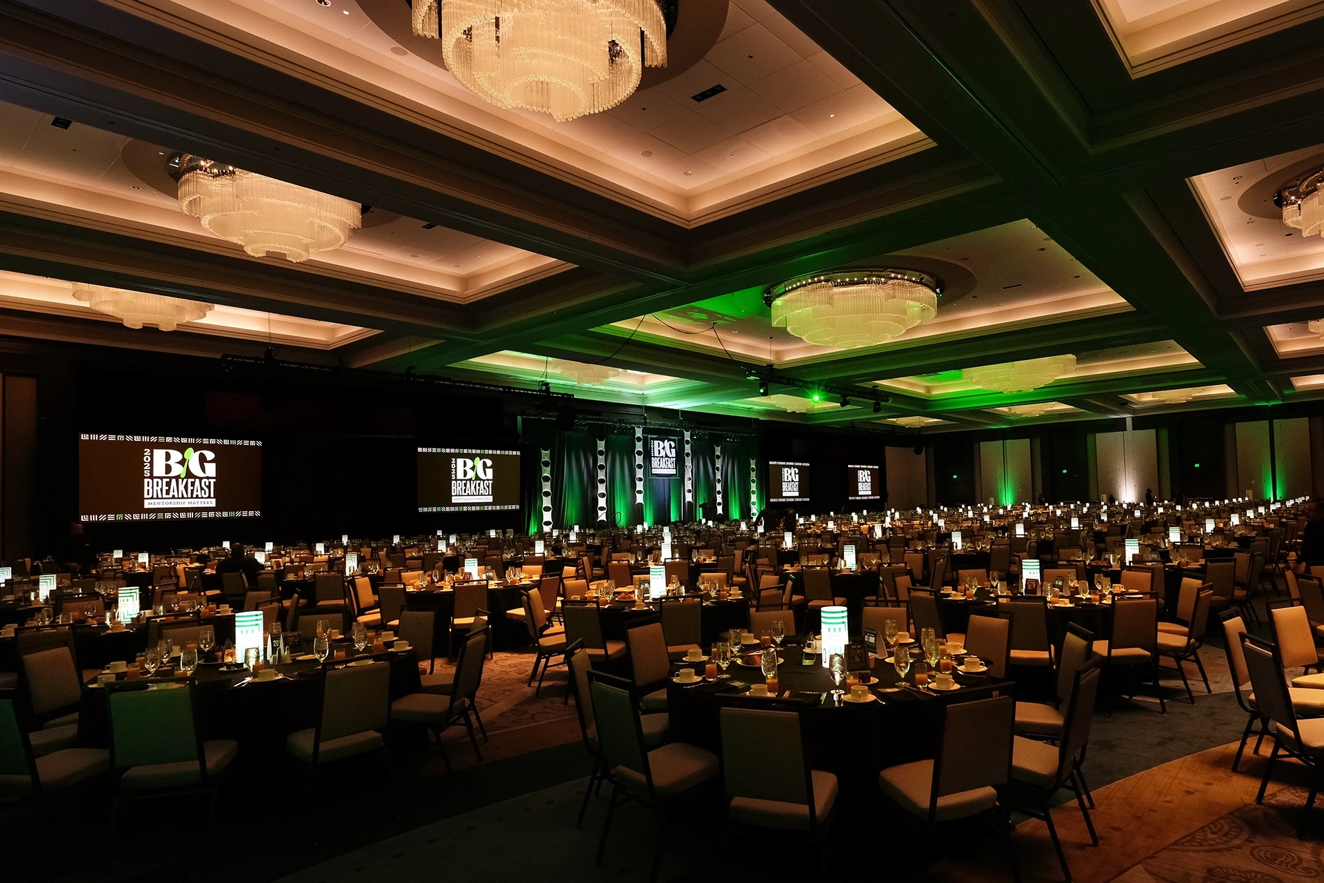 Round breakfast tables under a coffered ceiling and chandeliers with green uplighting at the Big Breakfast, Nashville — event production by UpLight Technologies
