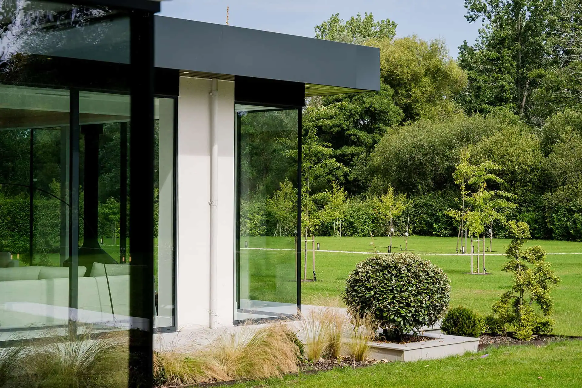 A modern glass structure with a black roof stands beside neatly trimmed greenery and landscaping.