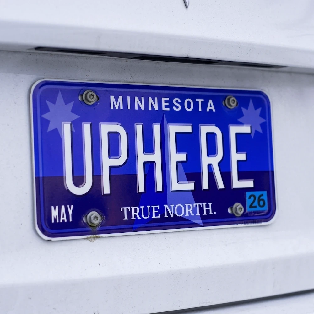 Close up photo of a blue Minnesota state license plate with the text "UPHERE" on a white car