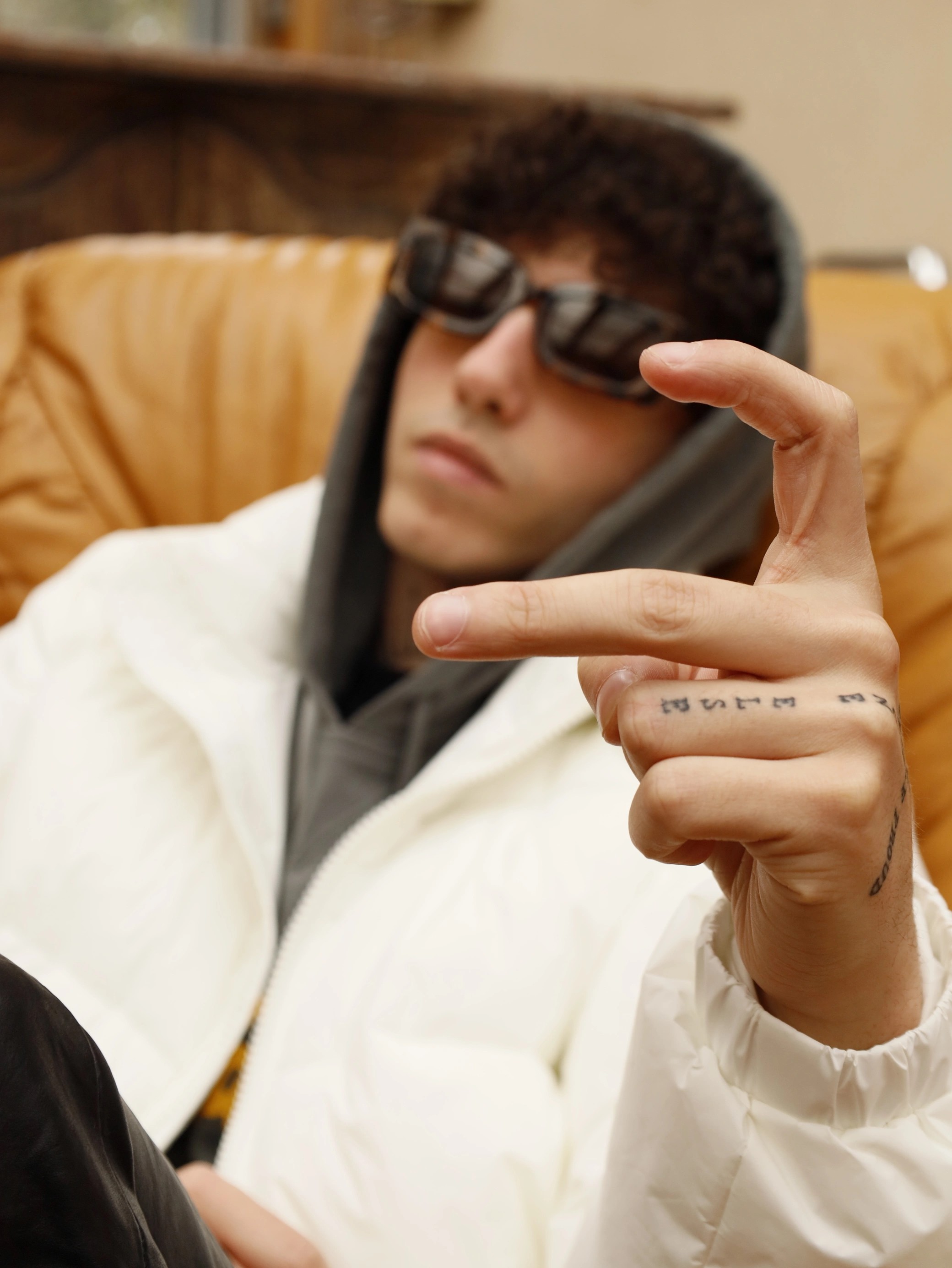 Person in a white jacket and sunglasses reclines on a tan chair, wearing a hoodie. They gesture with their fingers, displaying tattoos on one hand.