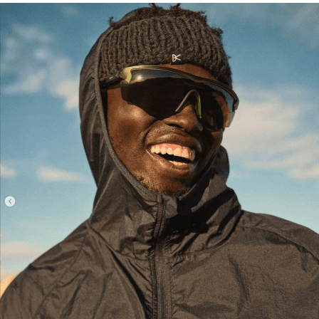 Smiling Black man wearing a beanie, sunglasses, and a hooded jacket, against a light blue sky.