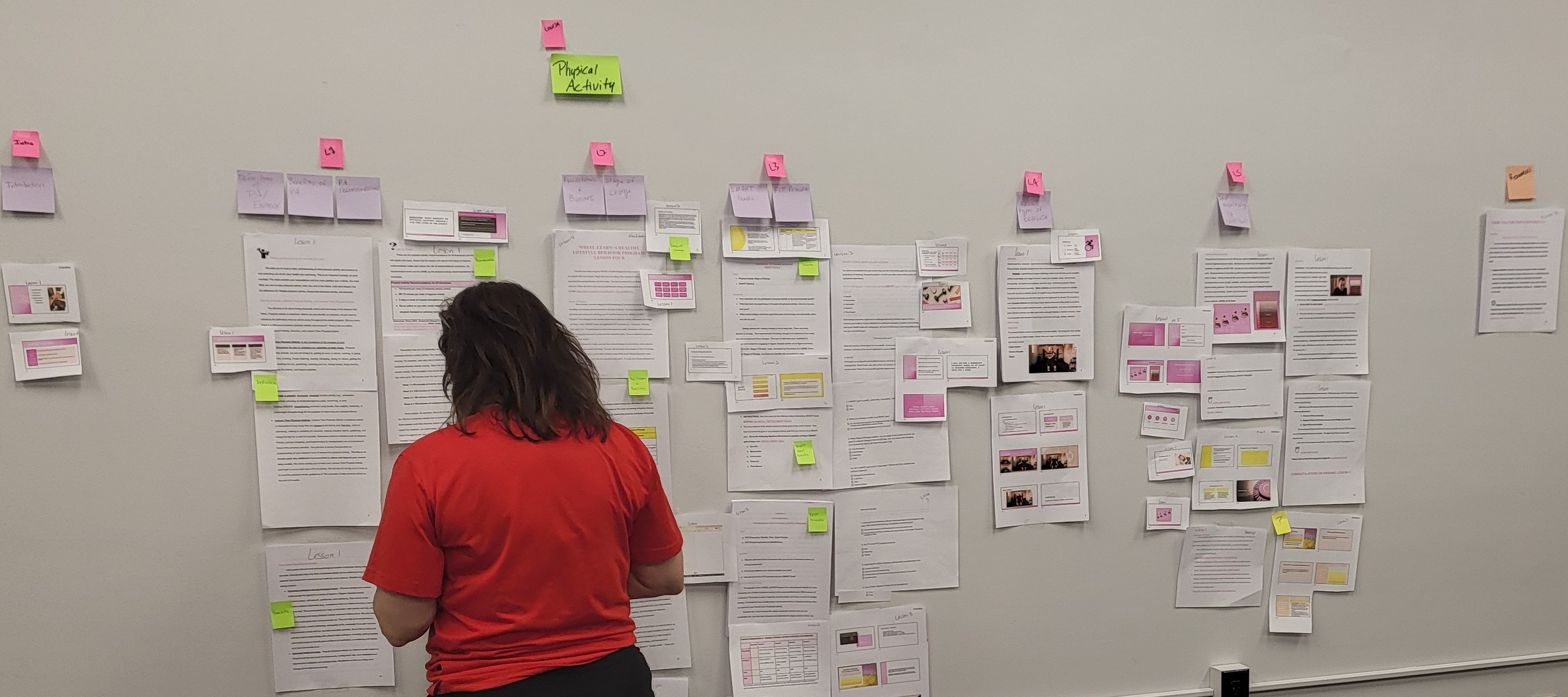 A woman in a red shirt stands in front of a wall covered in paper and sticky notes.