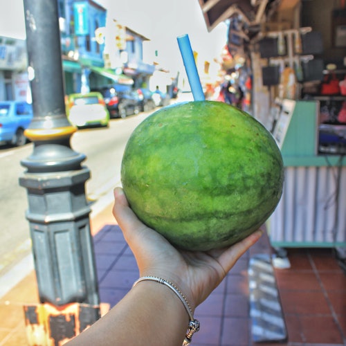 A hand holds a whole watermelon with a blue straw inserted, with a street and shops in the background.