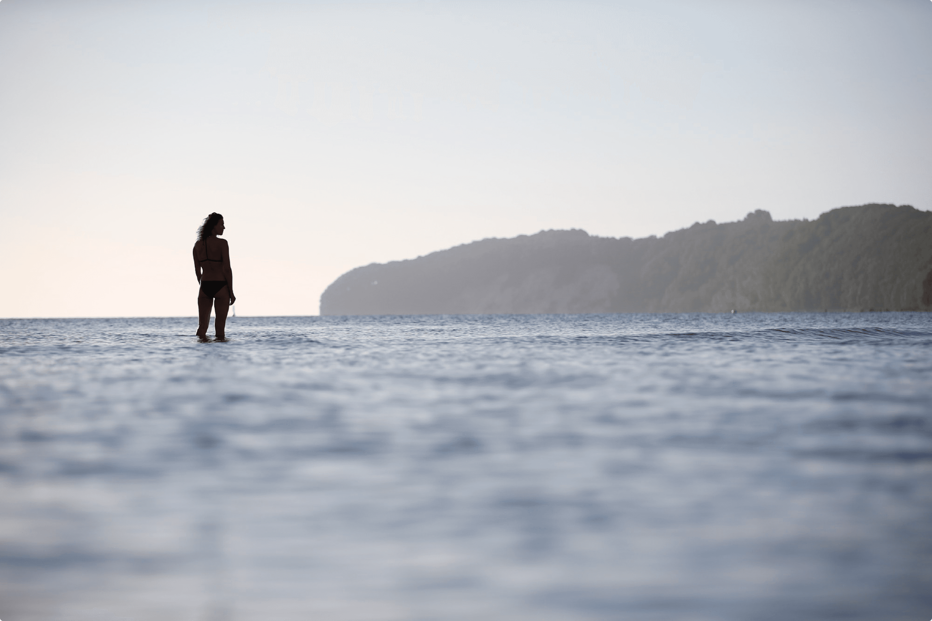 Woman in bathing suit stands in shallow water, looking out at calm sea.