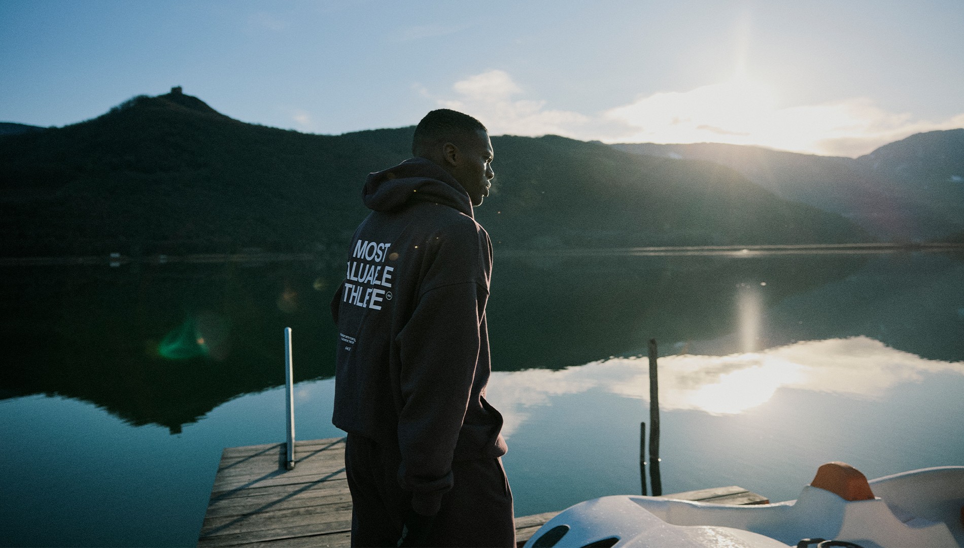 A man in a hoodie stands on a dock by a calm lake, with mountains and the sun low in the sky—a tranquil moment captured through oace-production-photography.