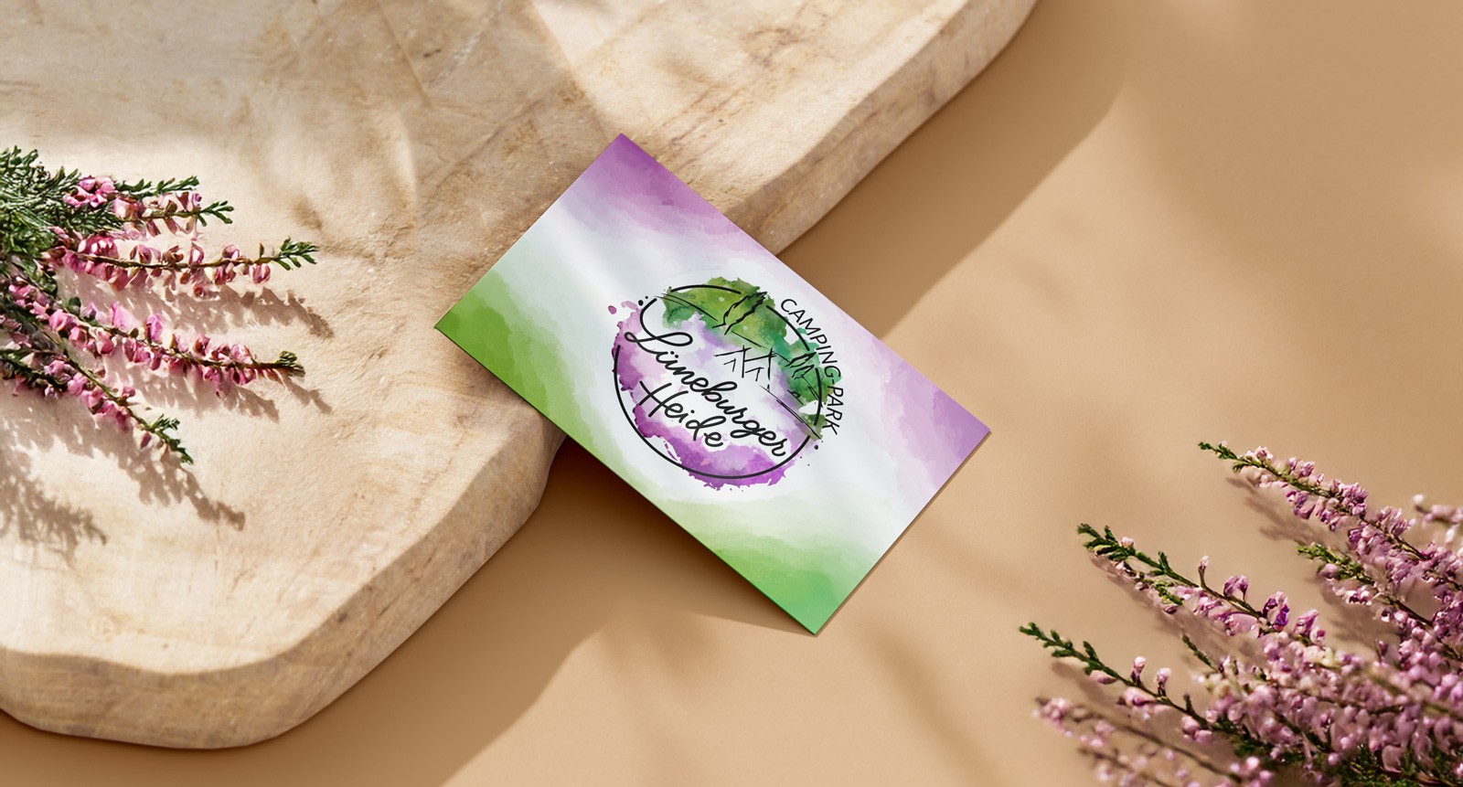 Visitenkarte für den Campingpark Lüneburger Heide mit mittig platziertem Logo und sanftem Aquarelldesign in den Hauptfarben Grün und Lila. Die Karte liegt auf beigem Untergrund mit Holzbrett, umrahmt von Heidezweigen in den Bild-Ecken.