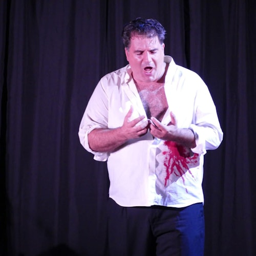 Un homme portant une chemise blanche avec une tache rouge, peut-être de sang, sur l'abdomen, se tient devant un rideau noir, l'air inquiet.