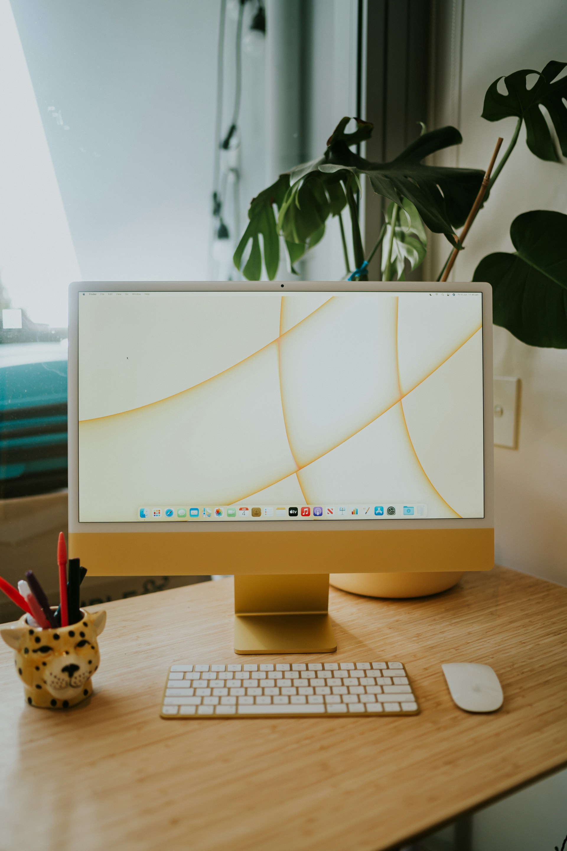 A yellow Apple iMac computer with a white keyboard and mouse sits on a wooden desk next to a potted plant and a leopard-shaped pen holder.