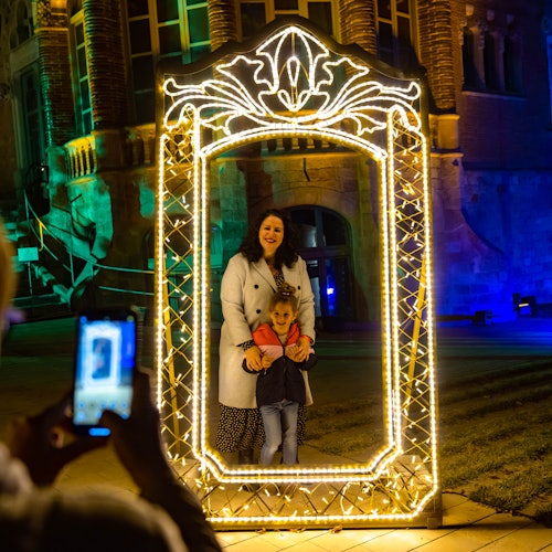 一位女性和孩子在一个大型发光框架内摆姿势,同时有人在夜晚拍摄发光建筑前的照片。