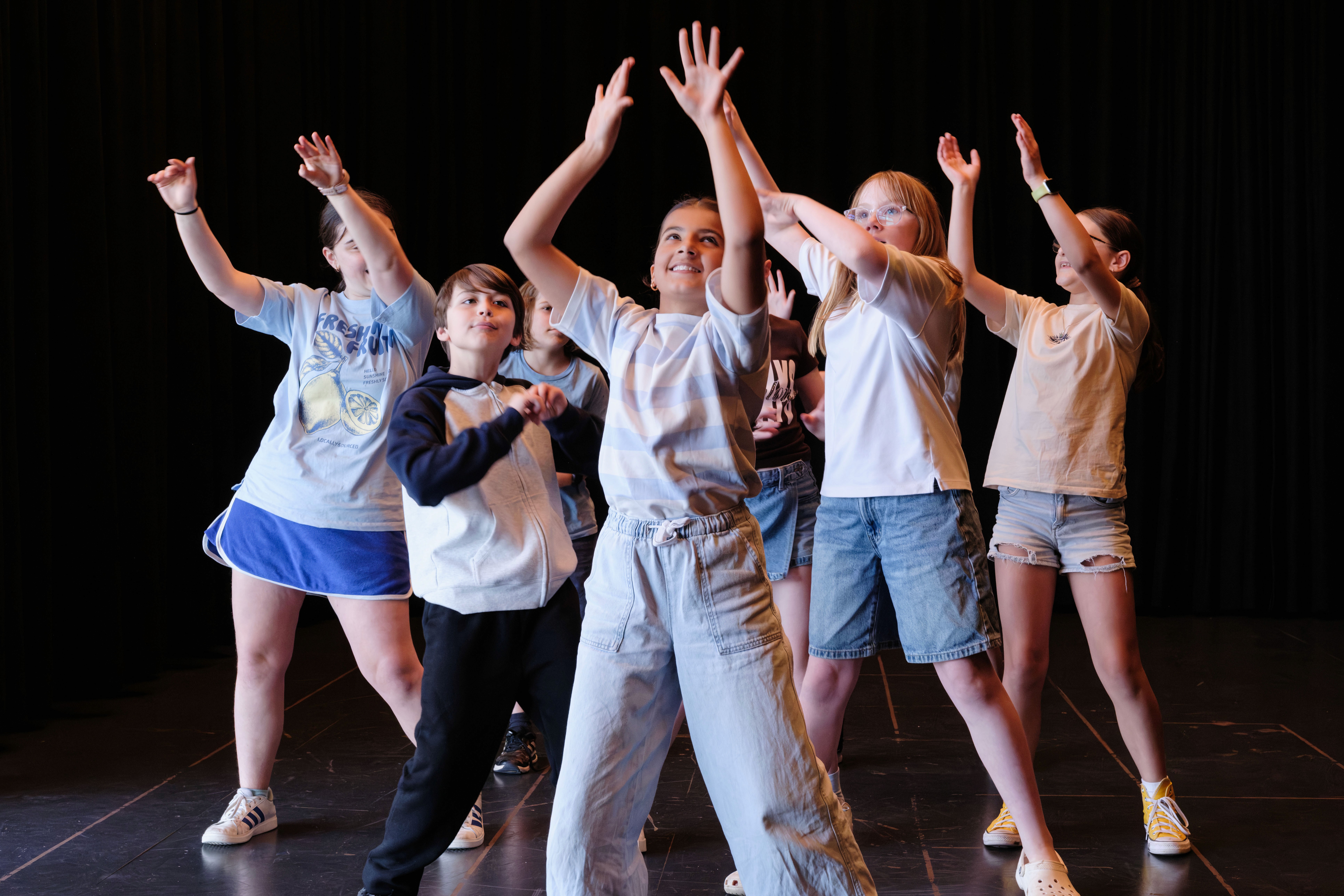 A group of young performers stand closely together in a rehearsal room, each extending an arm in a different direction as part of a movement exercise.