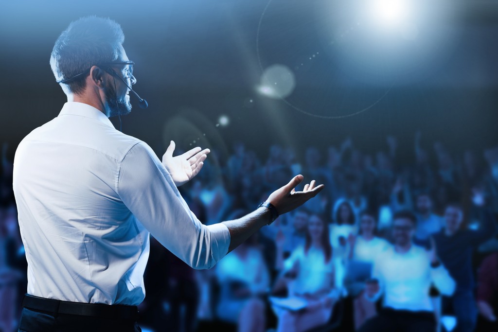 hero section background with plenty of space to the left out of focus; subject is a woman of color giving a speech from a podium with a smile and a laugh and confidence while others look on appreciatively