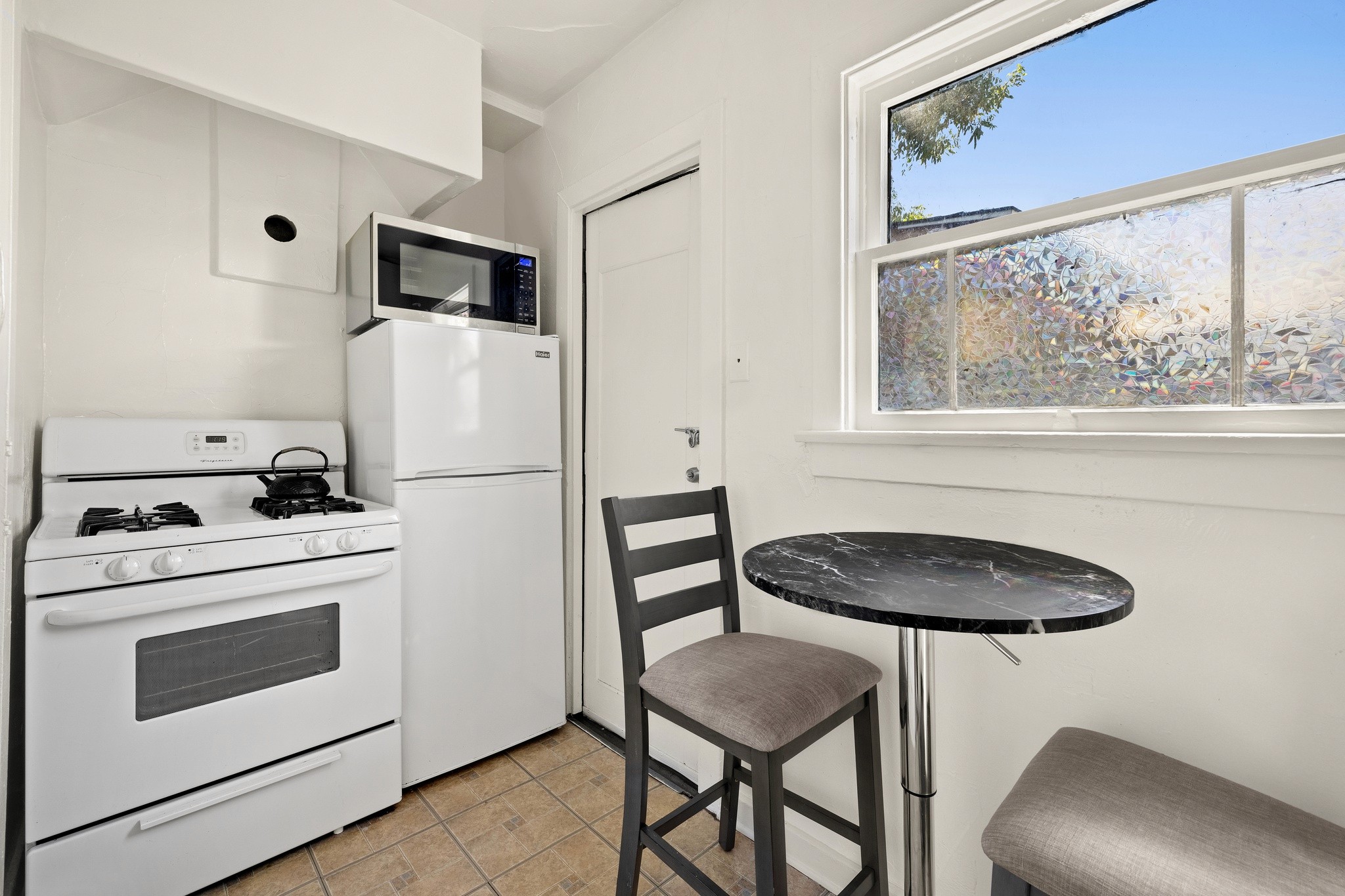 A small kitchen area at 3456 plata, featuring a stove, fridge, and a round table with chairs by a window. Bright and airy space.