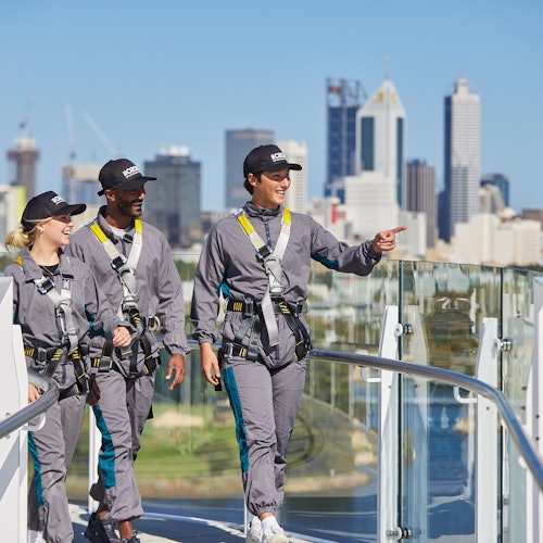 Three people in gray jumpsuits and harnesses walk along a high platform with a city skyline in the background. One person points ahead.