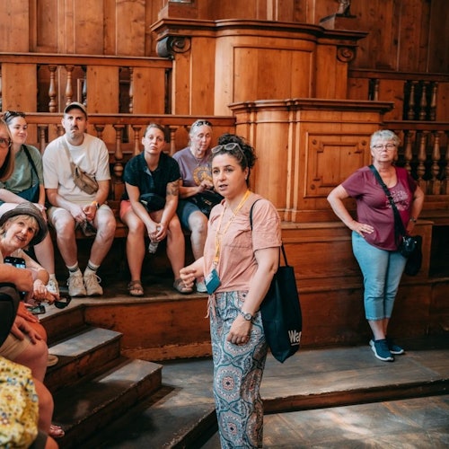 אישה המחזיקה תג מדריך מדברת לקבוצת סיור ישובה בתוך מבנה היסטורי מעץ.