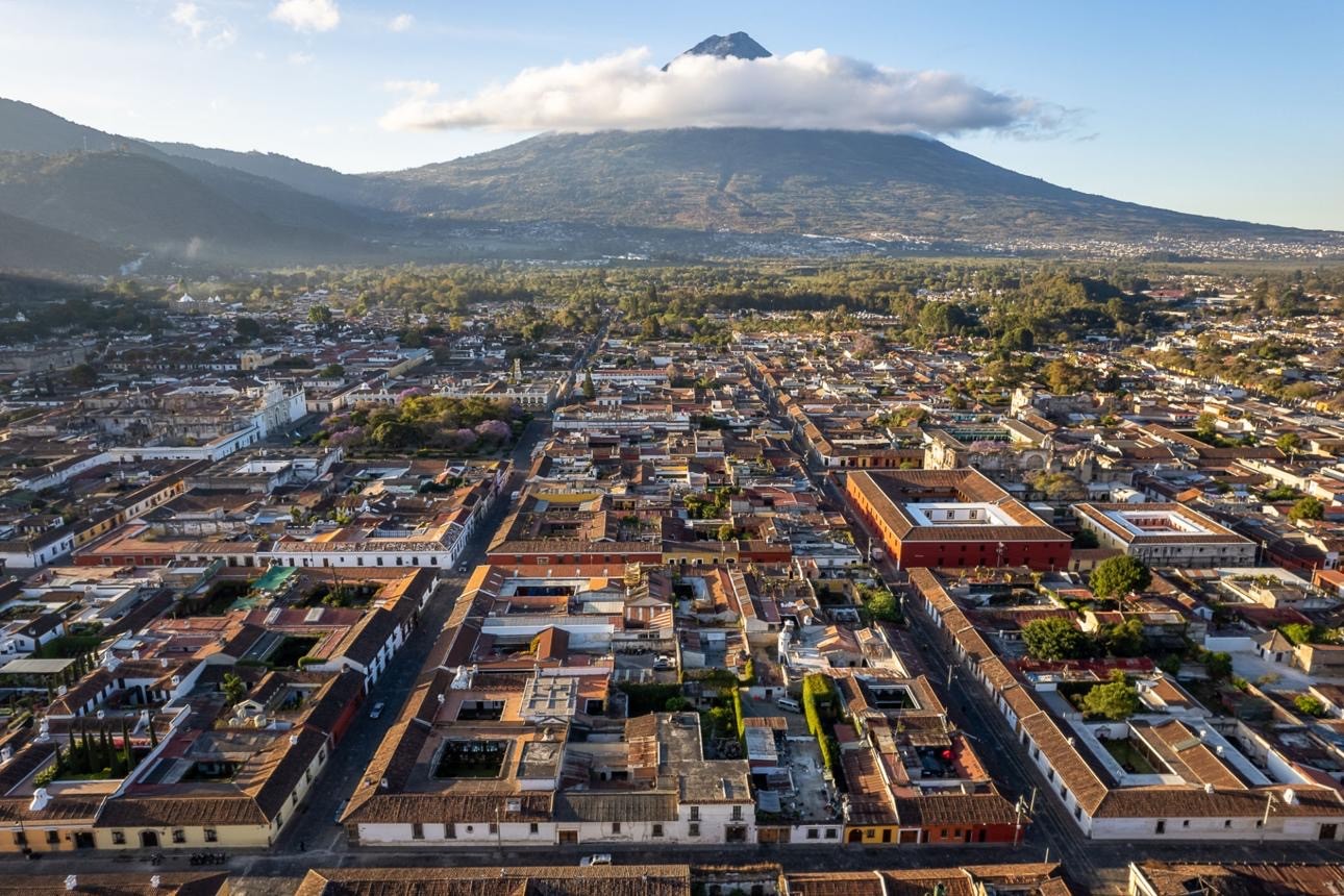 Aerial view of a vibrant townscape with rows of colorful, tiled-roof buildings nestled against a lush, mountainous backdrop, dominated by a towering volcano under a partly cloudy sky.
