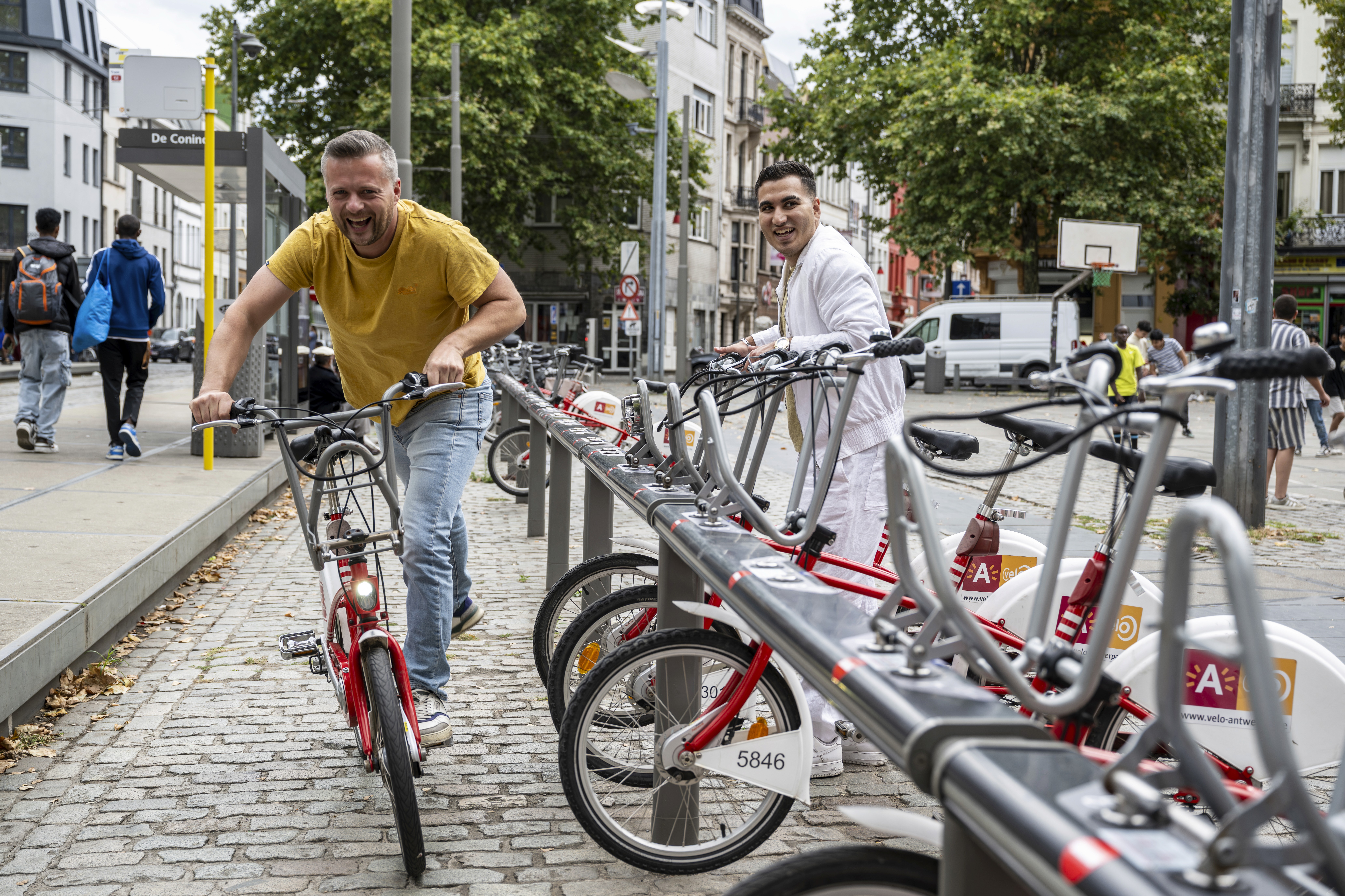 Two women on bicycles smile and chat while riding on a tree-lined path. One wears a floral dress, the other a white jacket and pants.