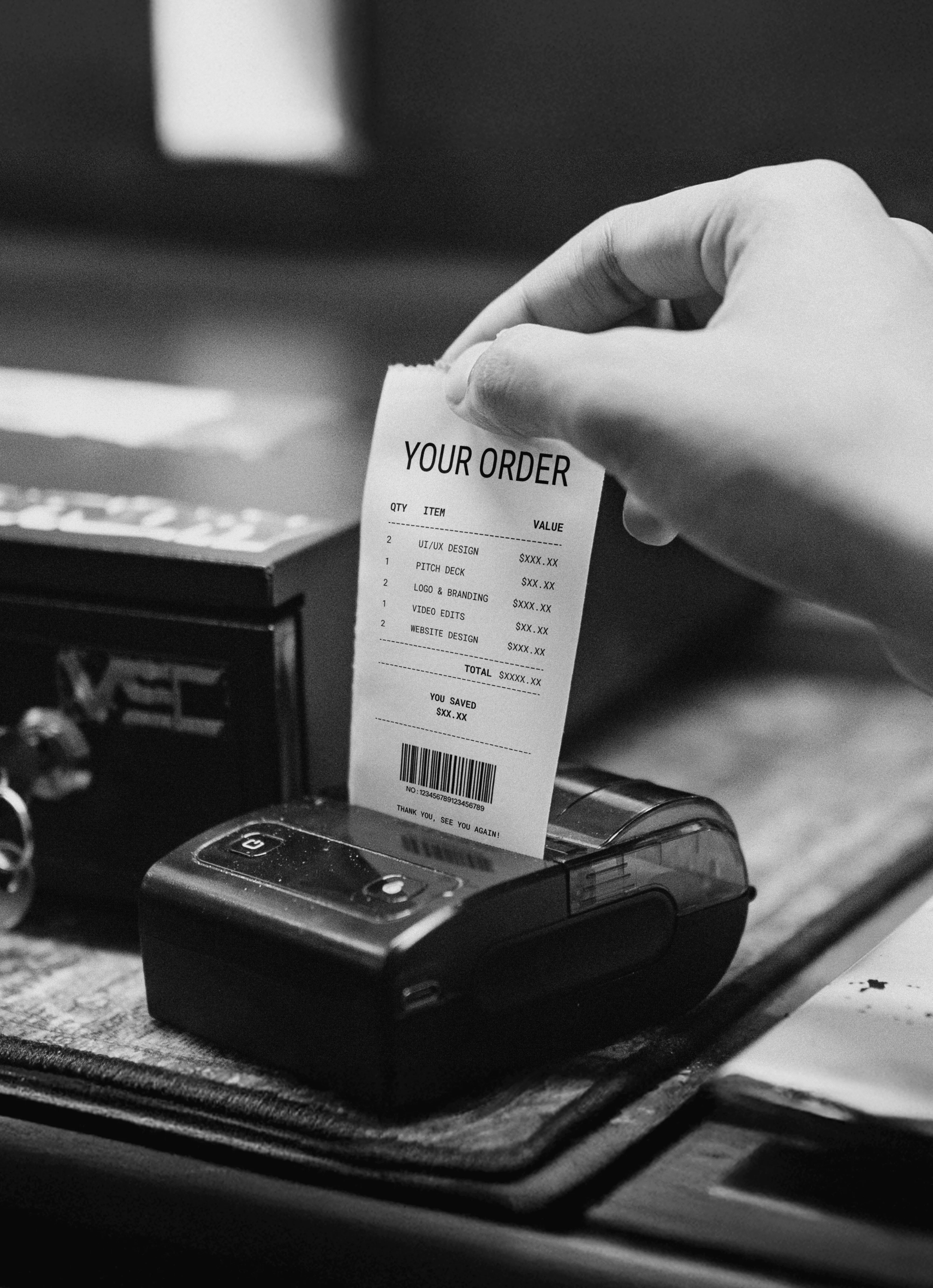 A black and white photo of a hand pulling a service receipt from a small printer, listing items like "UI/UX DESIGN" and "LOGO & BRANDING".
