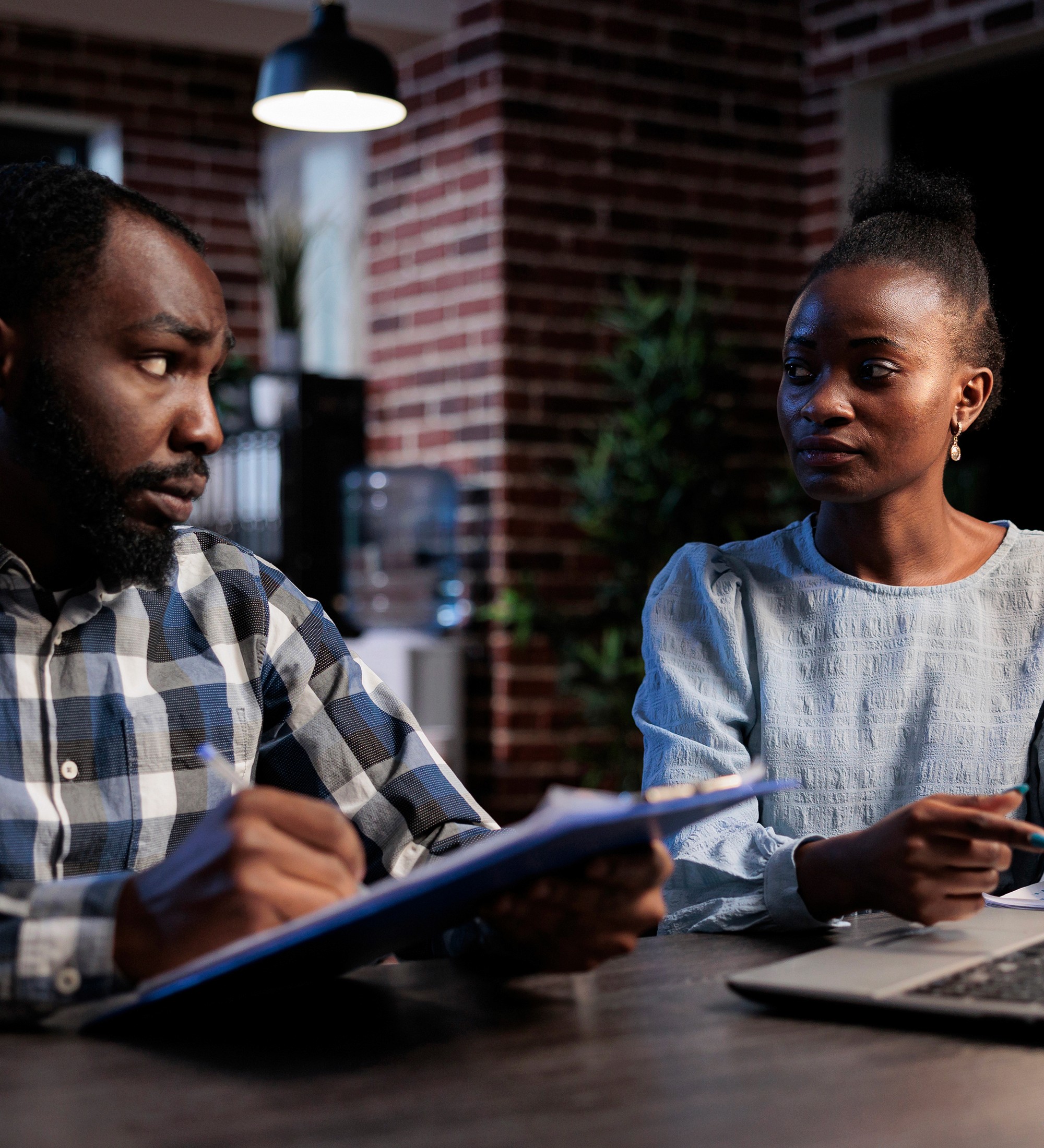 Two professionals sit at a table with exposed brick walls and industrial lighting in the background. One person in a plaid shirt is writing on a clipboard while the other in light-colored clothing appears to be engaged in conversation.