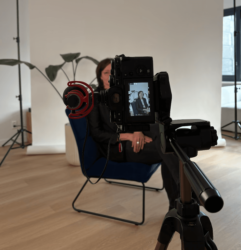A woman in a teal dress sits in a dimly lit room, focused on her smartphone. A laptop and notepad are on the glass table, suggesting a working environment.