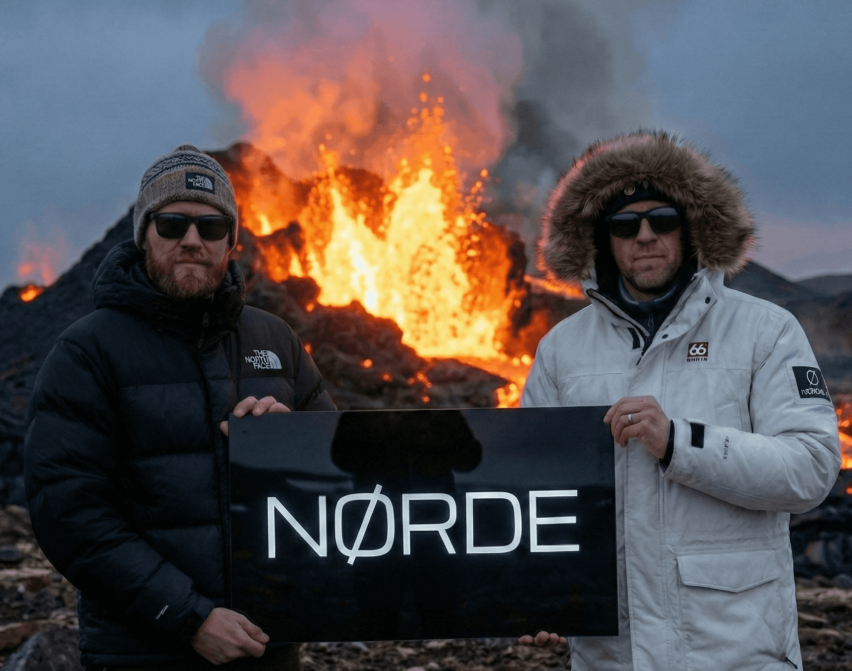 Two men in winter jackets and sunglasses standing in front of an active volcanic eruption with flowing lava, holding a black sign together.