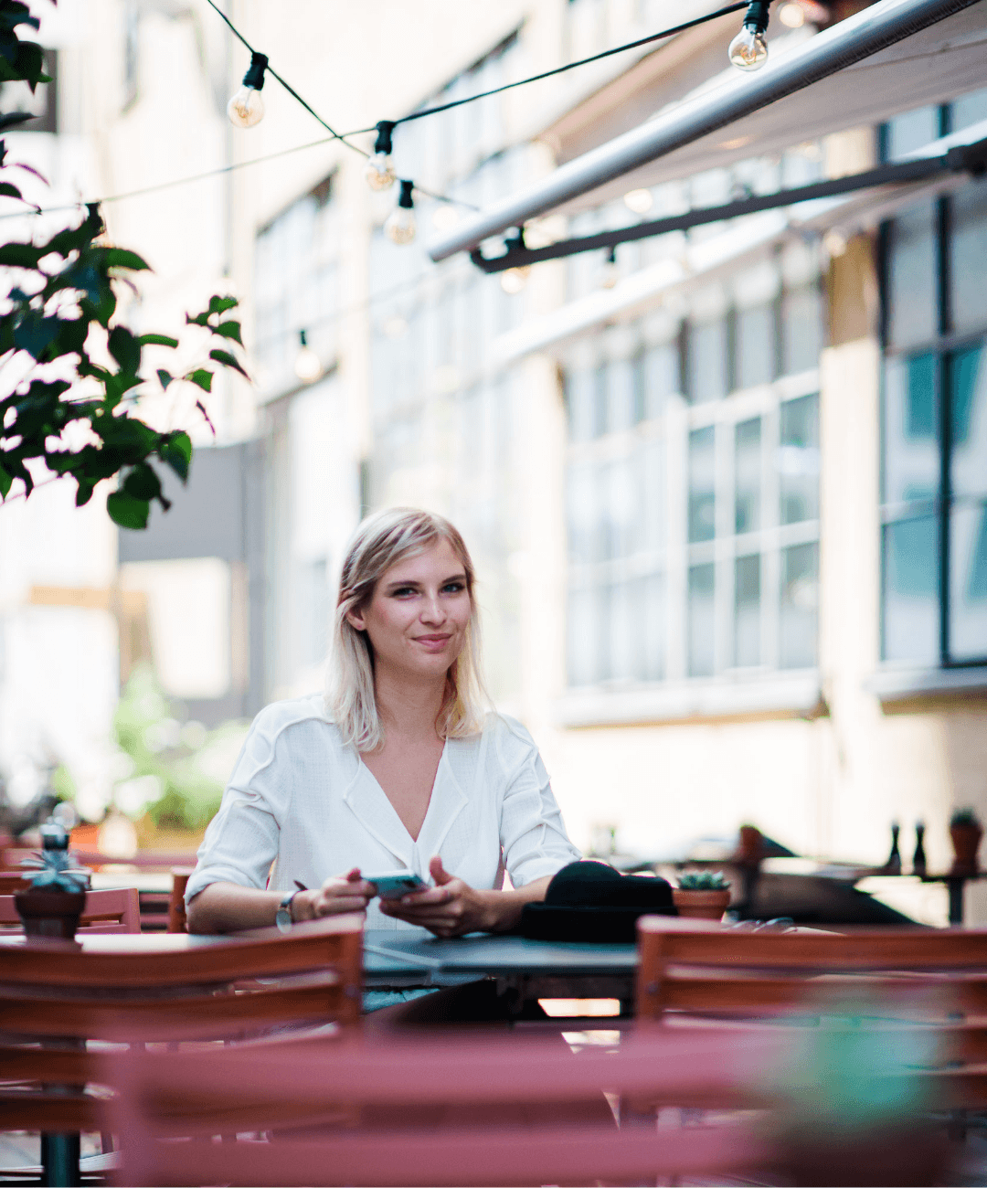 Blonde Frau in weisser Bluse sitzt lächelnd an einem Tisch im Freien. Sie hält ein Smartphone in den Händen. Im Hintergrund sind Gebäude und Lichterketten zu sehen, die eine gemütliche Atmosphäre schaffen.