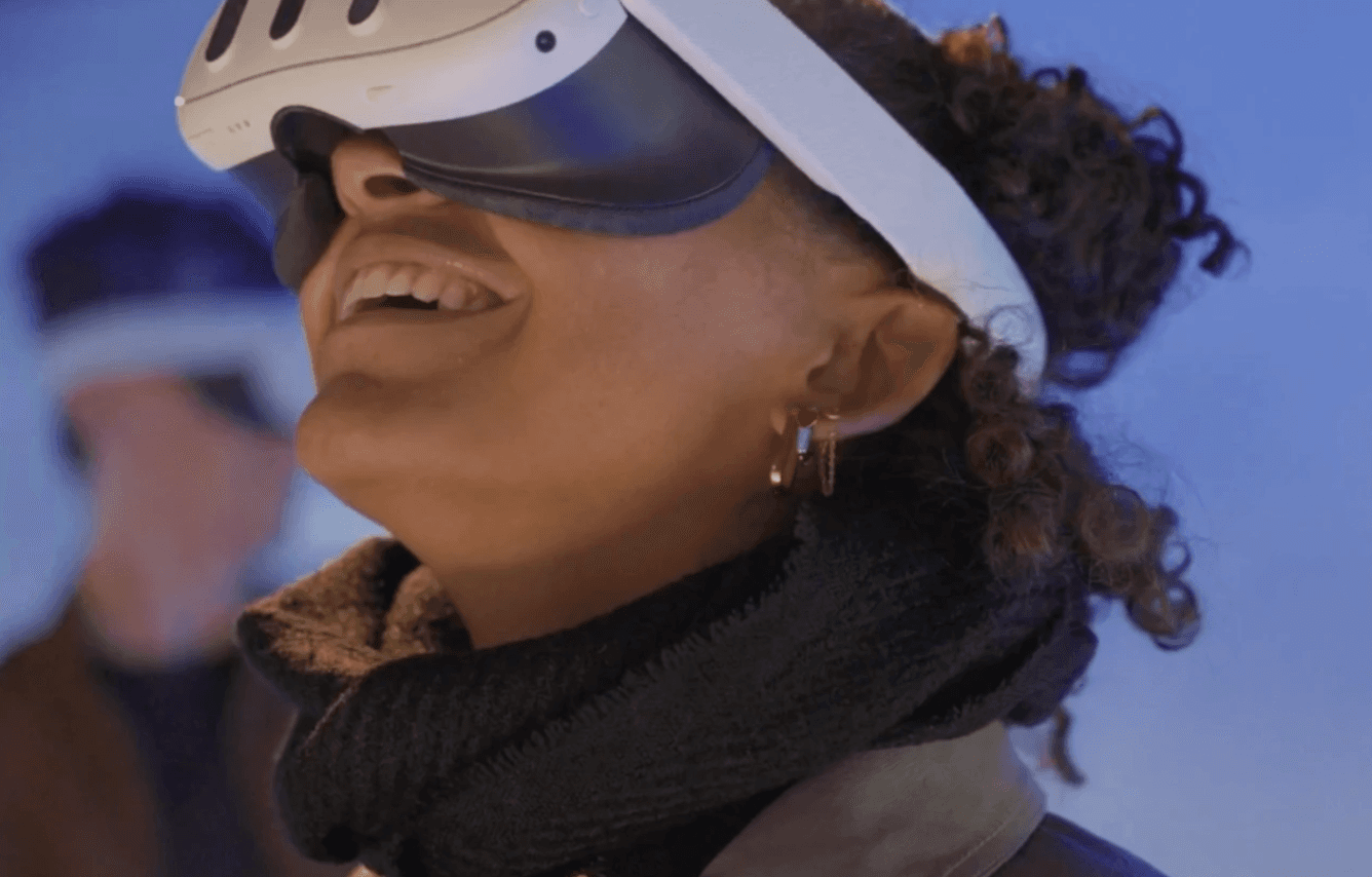 A man wearing glasses and a blazer working at a desk, writing in a notebook beside a computer monitor in a modern office.
