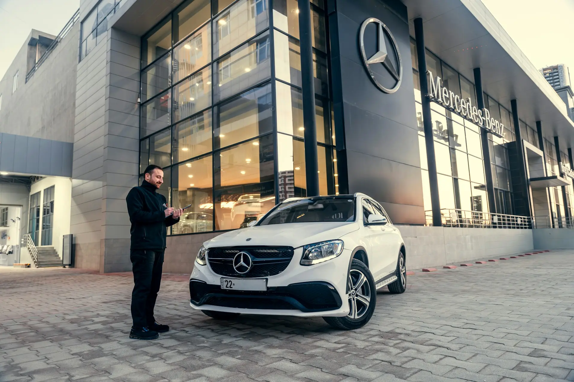 Car dealership exterior with a man standing next to a white SUV in front of a modern glass showroom building.