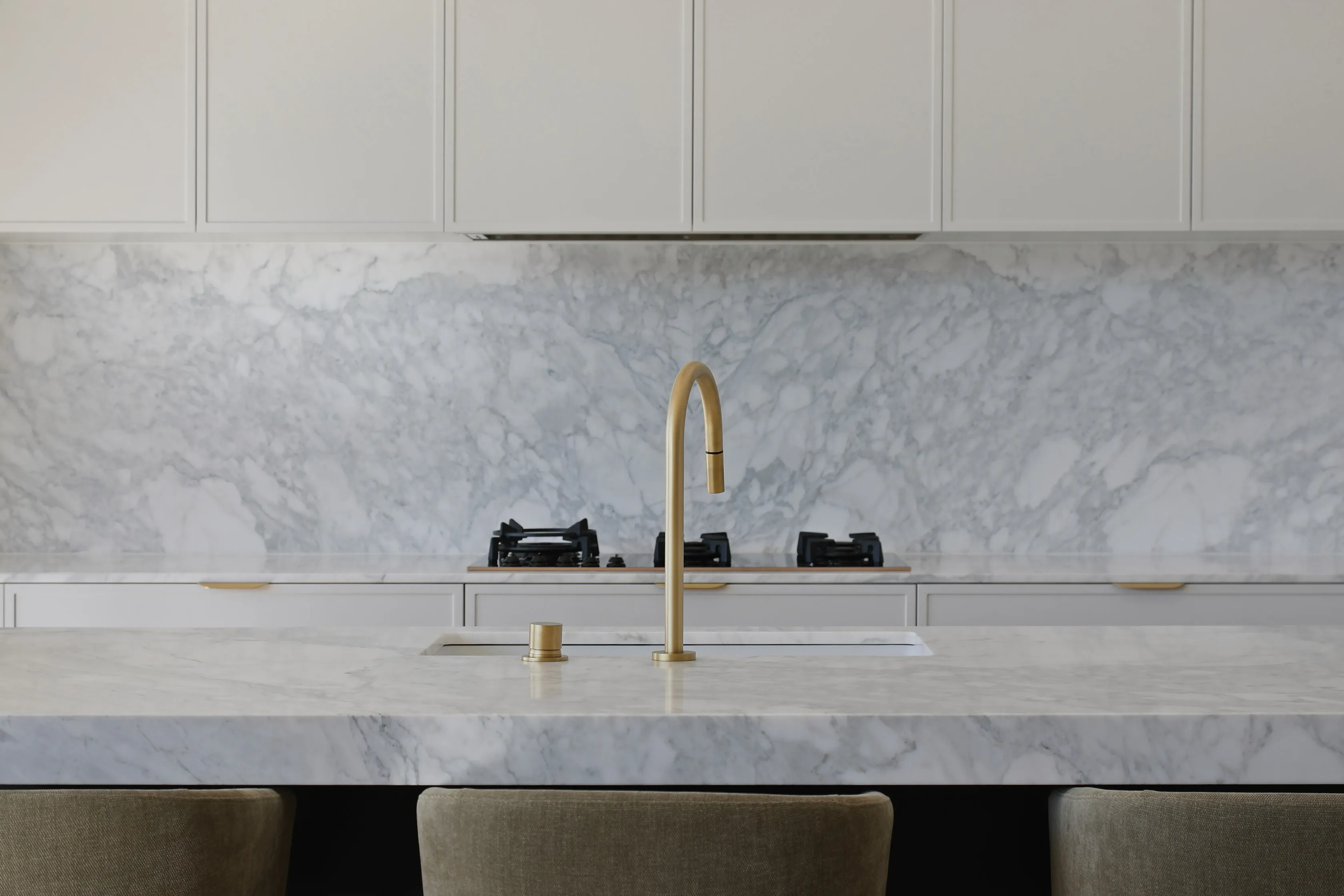 Kitchen detail with a brass gooseneck tap over an undermount sink and a black gas cooktop set in a Calacatta marble benchtop, against a matching marble splashback and white cabinetry