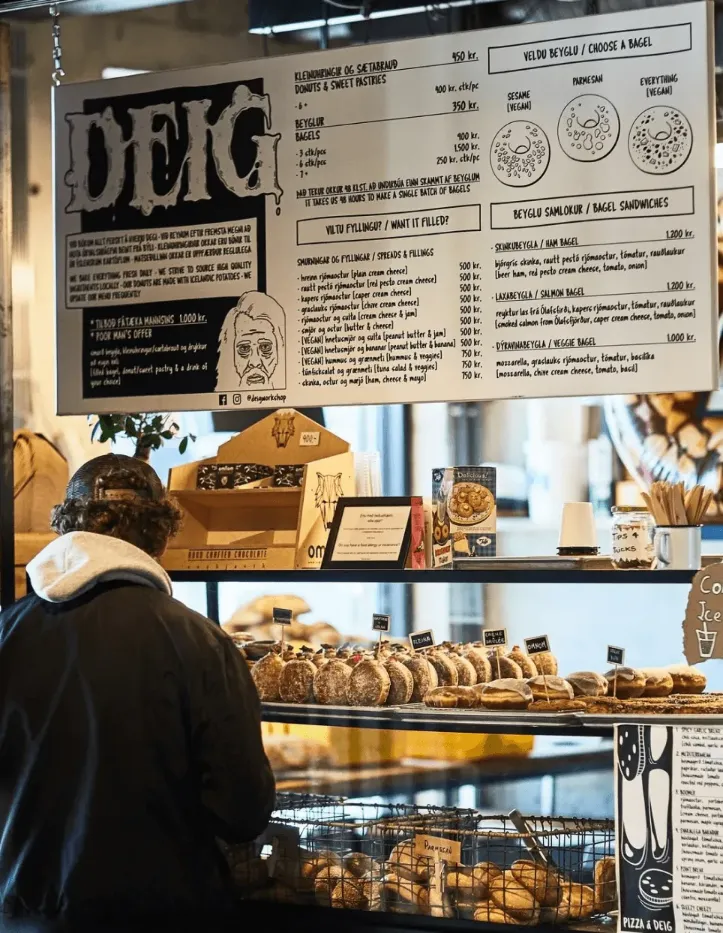 Interior of a DEIG bakery counter with a large illustrated menu, shelves of pastries and bagels, and a customer waiting at the counter.