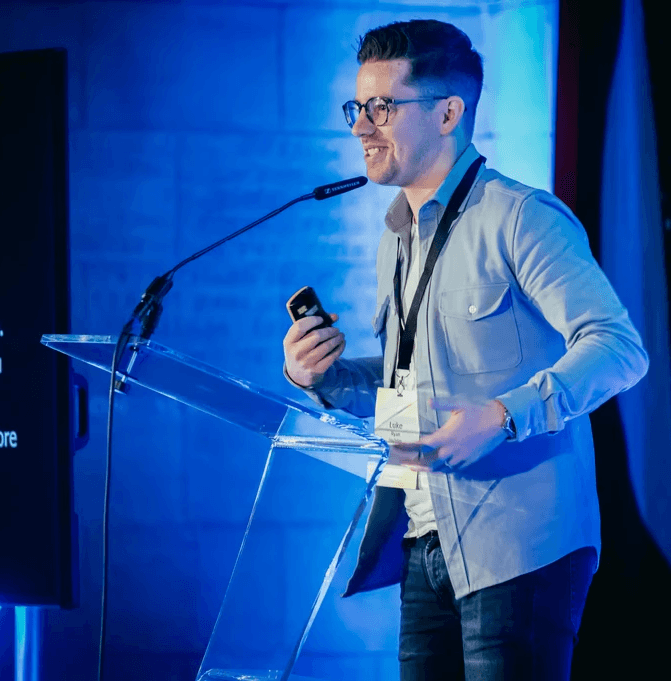 A person stands at a clear lectern on a stage, speaking into a microphone while holding a small handheld device. A large screen to the left displays partially visible presentation text. The setting is lit with blue stage lighting, and the speaker wears a light button-up shirt and a lanyard with a name badge.