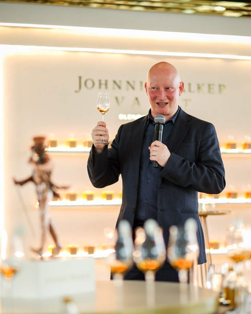 A presenter holds a glass of whisky while speaking into a microphone at a Johnnie Walker Vault event, with an array of whisky glasses in the foreground and a backdrop featuring the brand's logo.