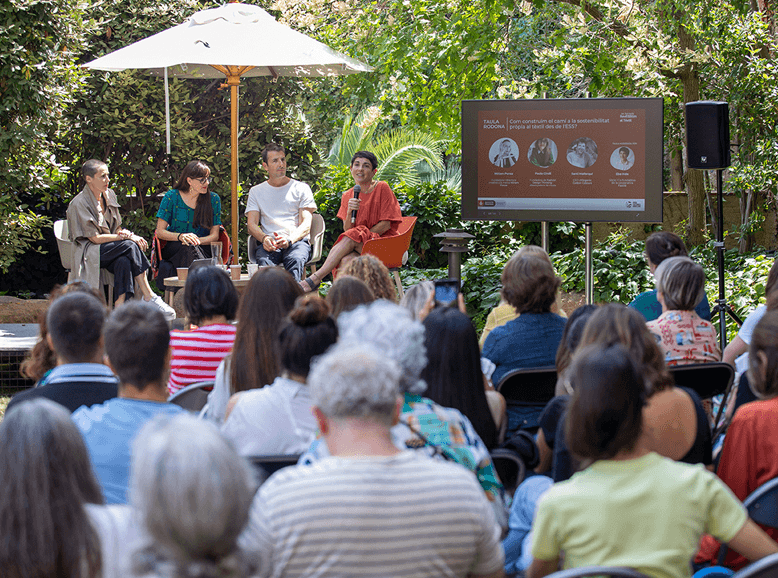 Panel discussion in a garden setting with four speakers on stage and an audience seated outdoors, facing a screen with visuals.